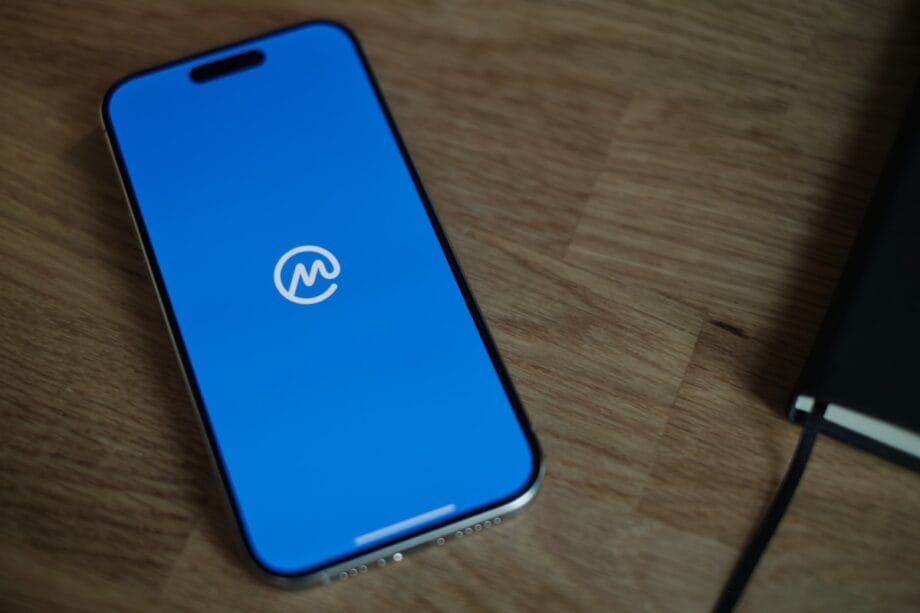 A smartphone with a blue screen displaying a white cryptocurrency app logo rests on a wooden surface next to a closed notebook.