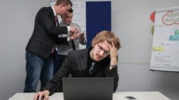 A man sits stressed at a laptop while two colleagues discuss papers in the background near a flip chart.