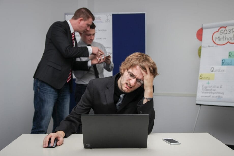 A man sits stressed at a laptop while two colleagues discuss papers in the background near a flip chart.