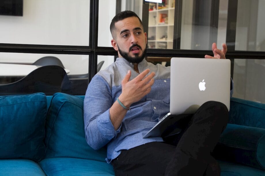 Man sitting on a blue couch using a laptop, gesturing with one hand while talking in a modern office setting.