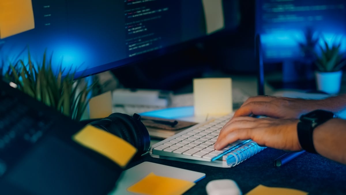 Person typing on a keyboard at a cluttered desk with sticky notes, computer monitor displaying code, and office items in dim lighting.