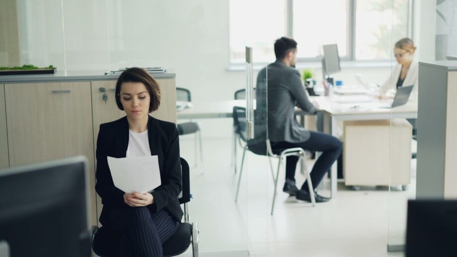 Woman in business attire sits reading a document in an office waiting area; two people work at desks behind glass in the background.