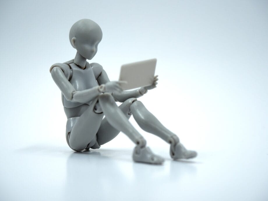 A gray humanoid mannequin sits on the ground holding a tablet against a plain white background.