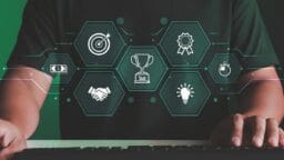 Person at keyboard with floating hexagon icons showing target, handshake, trophy, ribbon, clock, money, and lightbulb symbols.