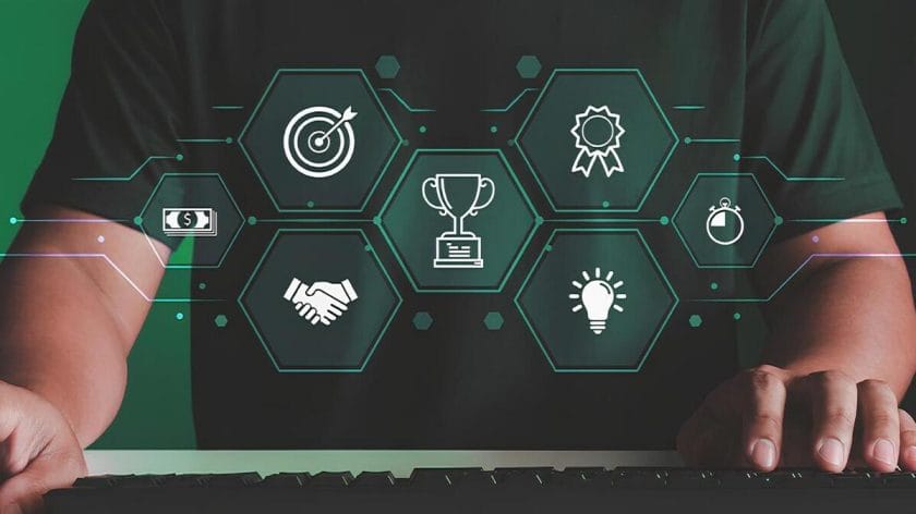 Person at keyboard with floating hexagon icons showing target, handshake, trophy, ribbon, clock, money, and lightbulb symbols.