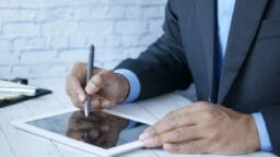 A person in a suit uses a stylus to write on a tablet placed on a white wooden table.