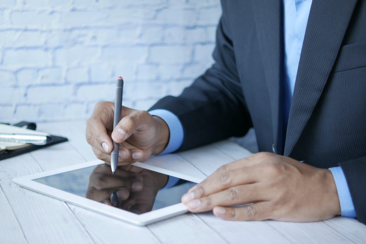 A person in a suit uses a stylus to write on a tablet placed on a white wooden table.