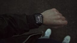 A person wearing a black smartwatch with an analog-style display, standing outdoors on a dark surface, wearing white shoes.