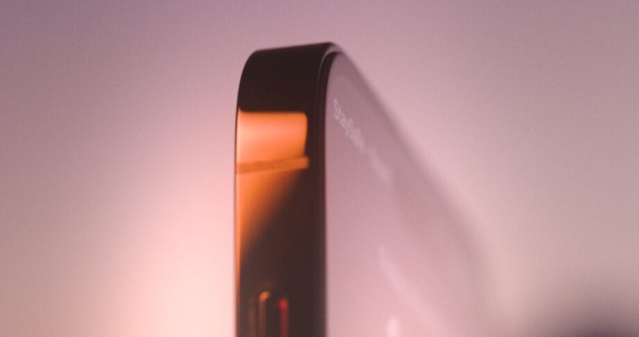 Close-up of the edge of a modern smartphone with a metallic frame and glass screen, illuminated by soft light.