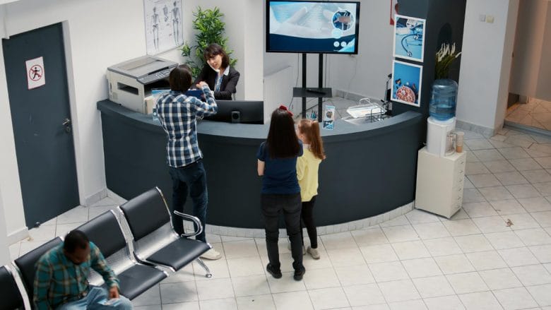A man checks in at a circular reception desk while a woman and child stand nearby in a medical waiting area.