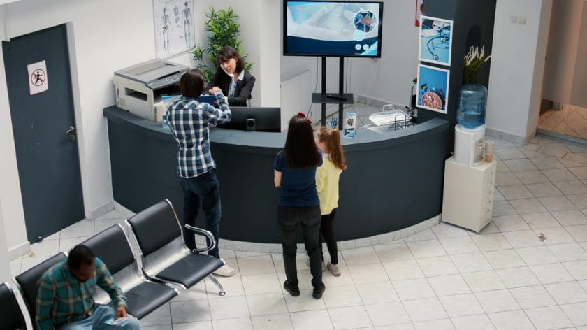 A man checks in at a circular reception desk while a woman and child stand nearby in a medical waiting area.