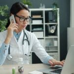 A doctor in a white coat uses a laptop and speaks on the phone at her desk, with medicine bottles and office shelves in the background.