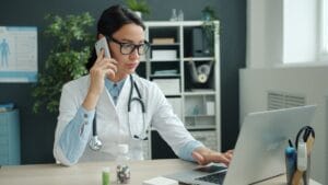 A doctor in a white coat uses a laptop and speaks on the phone at her desk, with medicine bottles and office shelves in the background.