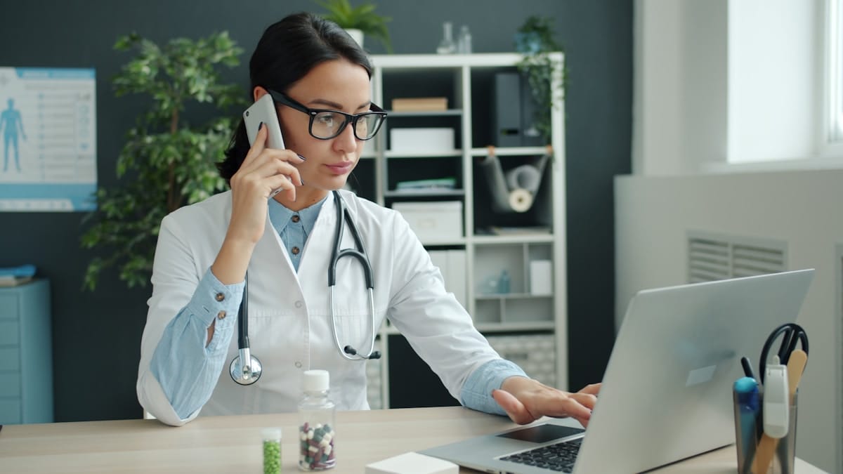 A doctor in a white coat uses a laptop and speaks on the phone at her desk, with medicine bottles and office shelves in the background.