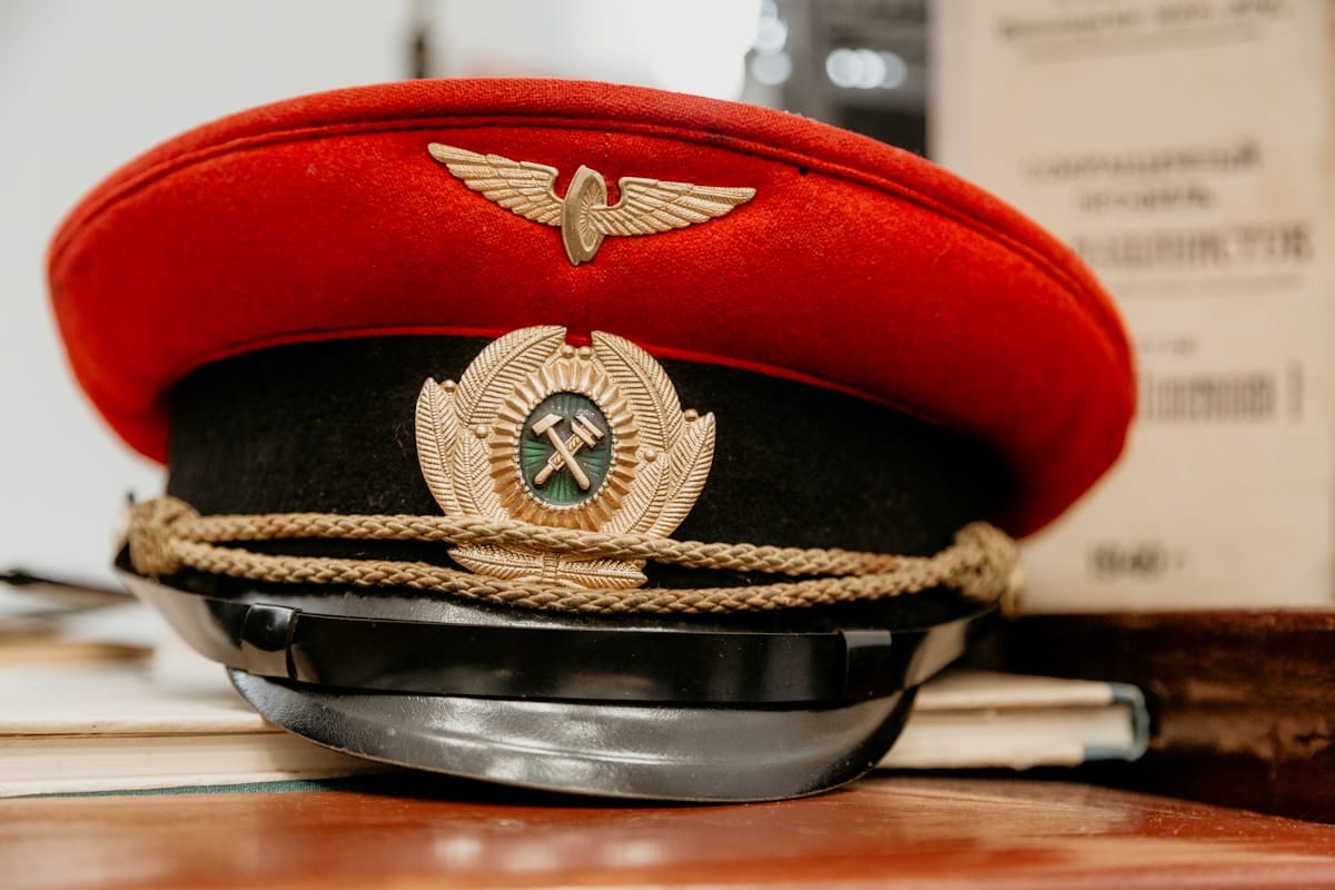 A red and black peaked cap with gold insignia, including wings and crossed hammers, rests on a stack of papers.