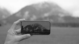 A hand holds a smartphone showing a clear view of a mountain, while the actual mountain in the background appears blurry and foggy.