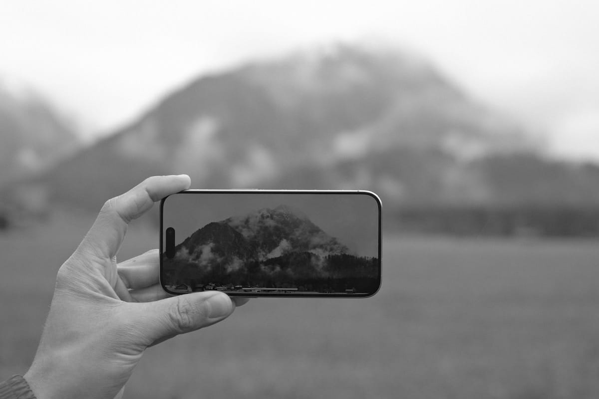 A hand holds a smartphone showing a clear view of a mountain, while the actual mountain in the background appears blurry and foggy.