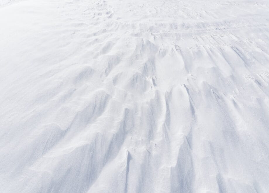 Close-up view of wind-sculpted snow drifts forming textured patterns on a flat, snow-covered surface.