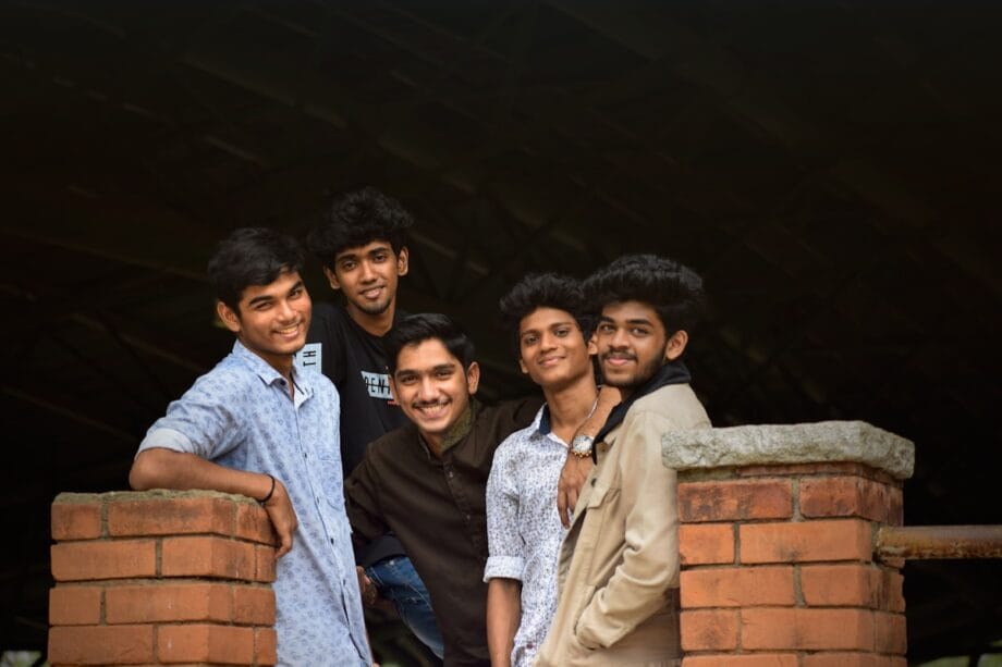 Five young men standing together and smiling in front of a brick wall with a dark background.