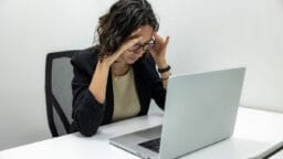 A woman sits at a desk, looking stressed as she holds her head with both hands in front of an open laptop.