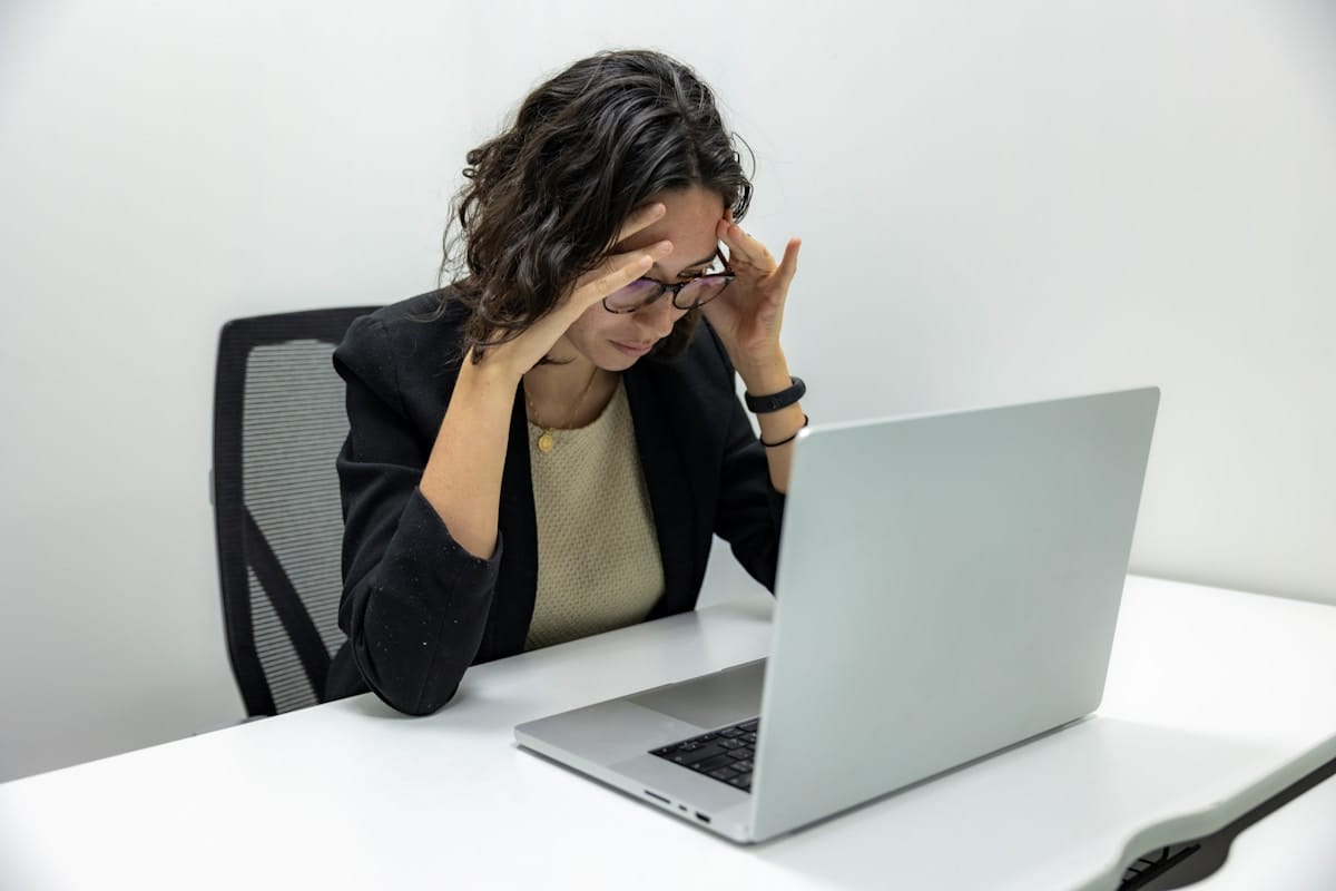 A woman sits at a desk, looking stressed as she holds her head with both hands in front of an open laptop.