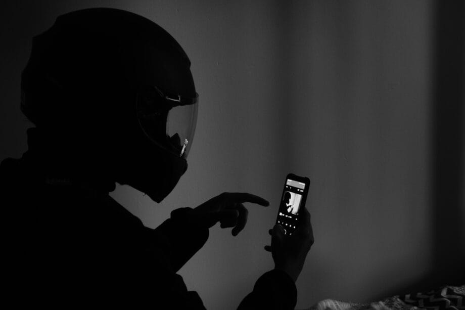 A person wearing a motorcycle helmet sits indoors, using a smartphone with one hand while pointing at the screen with the other.
