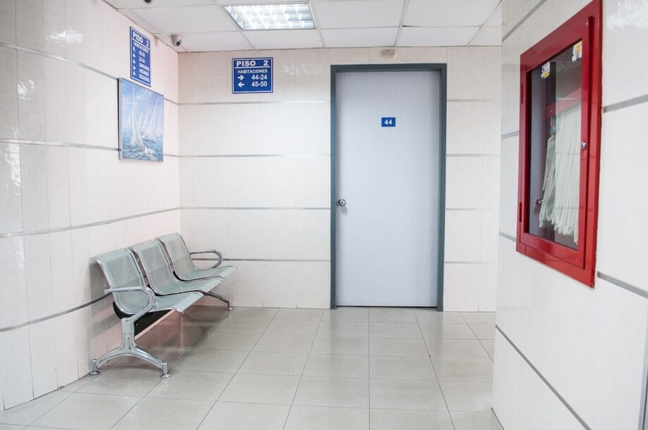 Empty waiting area with metal chairs, a closed door marked 44, and a red emergency equipment box on a tiled wall.