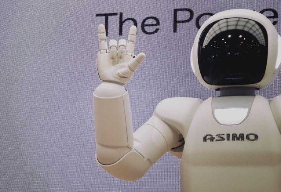 A white humanoid robot labeled ASIMO raises its hand, forming the I love you sign in sign language. Text is partially visible behind.