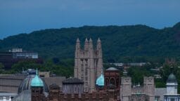 A cluster of historic stone buildings with towers and green domes, set against a backdrop of trees and a forested hill.