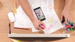 Person highlighting notes on paper with a smartphone, markers, and computer on a wooden desk, seen from above.