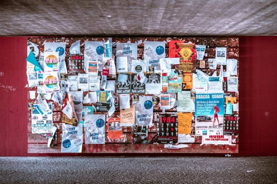 A bulletin board covered with various posters, flyers, and notices under a concrete ceiling and surrounded by a red wall.