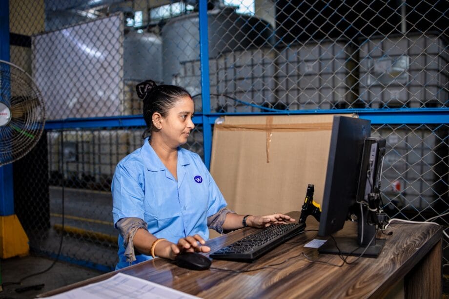 Woman in a blue uniform works on a desktop computer at a desk inside an industrial warehouse.
