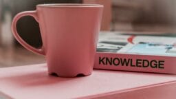 A pink mug sits on a pink surface next to a book titled KNOWLEDGE.