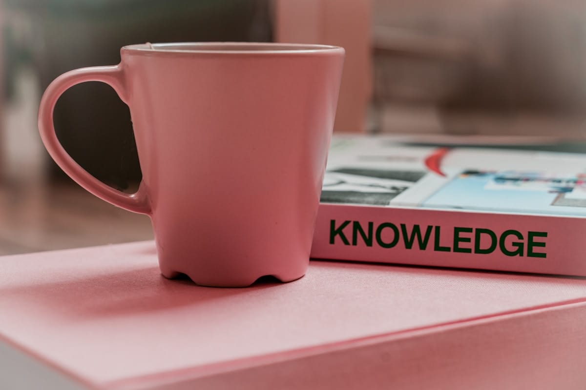 A pink mug sits on a pink surface next to a book titled KNOWLEDGE.