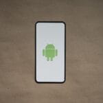 A smartphone displays the green Android robot logo on a white screen, placed on a plain brown surface.