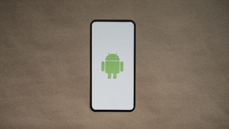 A smartphone displays the green Android robot logo on a white screen, placed on a plain brown surface.