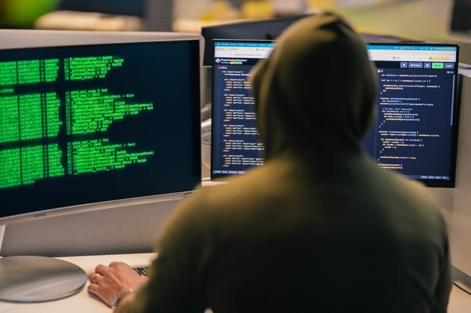 A person in a hoodie sits at a desk with two monitors displaying code and green text on dark backgrounds.