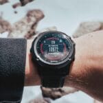 A person’s wrist wearing a digital watch displaying altitude at 10,972 feet, with a snowy, rocky background.