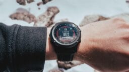 A person’s wrist wearing a digital watch displaying altitude at 10,972 feet, with a snowy, rocky background.