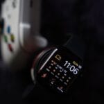 A smartwatch displaying weather, time, and date is shown next to part of a white video game controller on a dark surface.