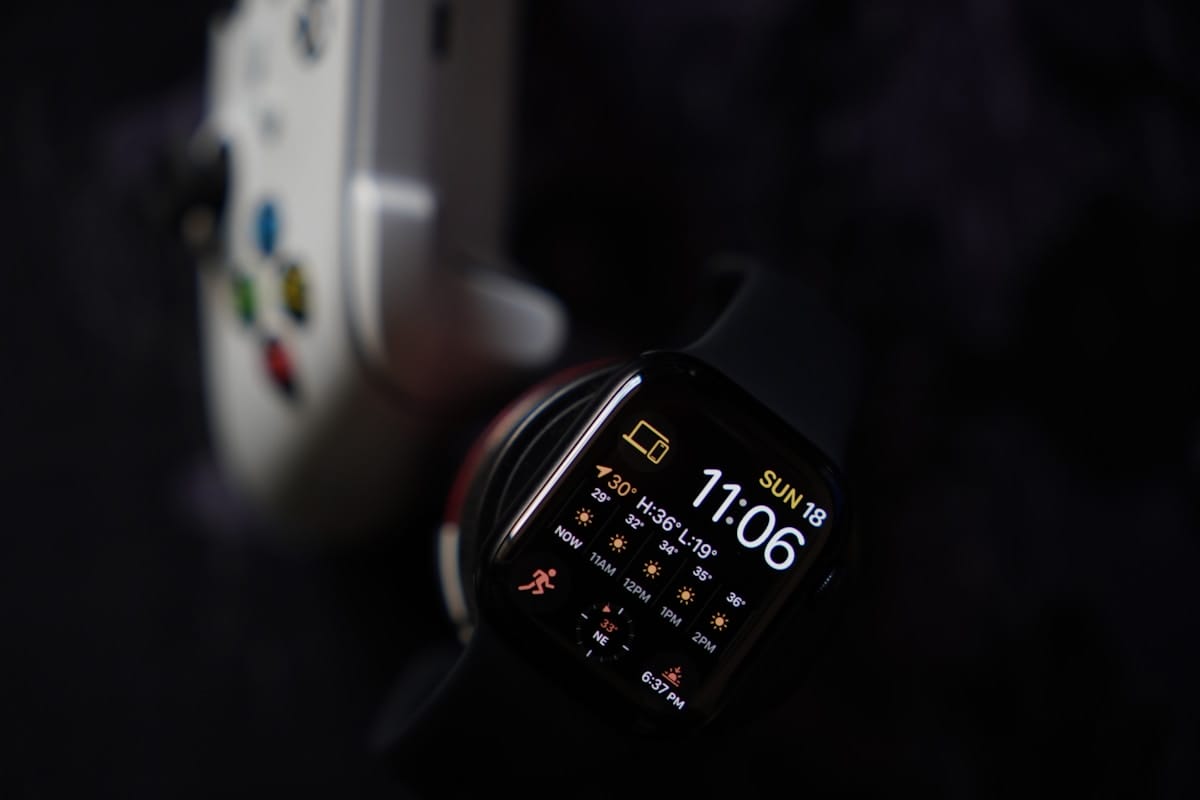 A smartwatch displaying weather, time, and date is shown next to part of a white video game controller on a dark surface.