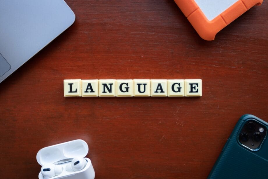 Letter tiles spelling LANGUAGE on a wooden table, surrounded by a laptop, tablet, wireless earbuds, and a smartphone.