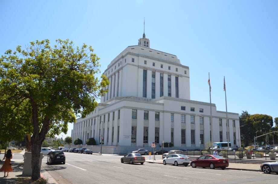Large white government building with columns, cars parked in front, and two flagpoles on a sunny day.