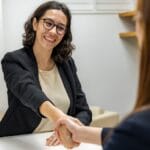 Two women sitting at a table shaking hands during a meeting or interview in an office setting.
