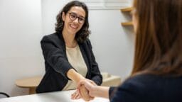 Two women sitting at a table shaking hands during a meeting or interview in an office setting.