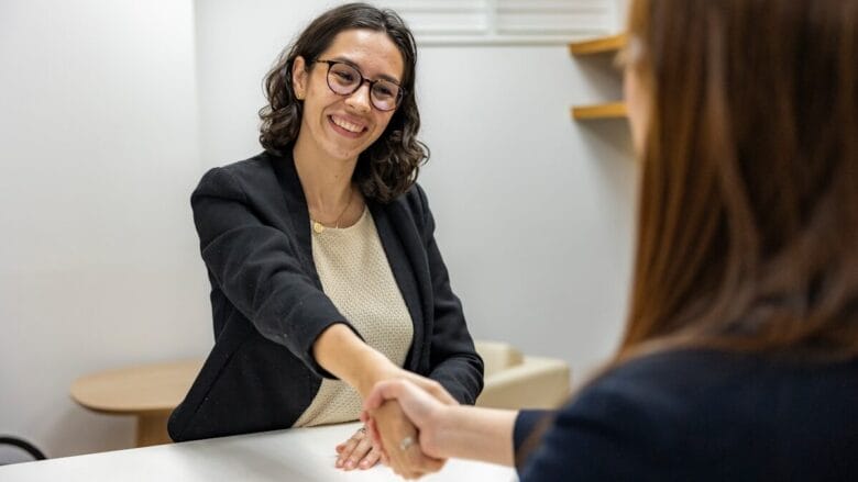 Two women sitting at a table shaking hands during a meeting or interview in an office setting.
