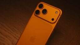 An orange smartphone with three rear cameras and a flash lies on a wooden surface, viewed from an angle.