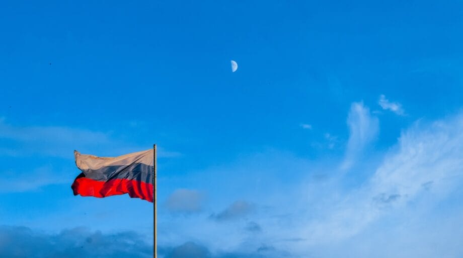 A Russian flag on a pole waves against a blue sky with scattered clouds and a visible moon.