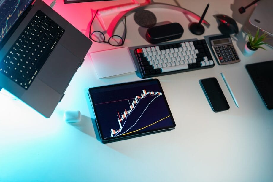 A tablet displaying a financial chart sits on a desk with a laptop, keyboard, calculator, phone, and office supplies.