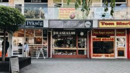 A row of storefronts with signs in Hungarian, featuring a bakery, a salon, and other shops on a pedestrian street.